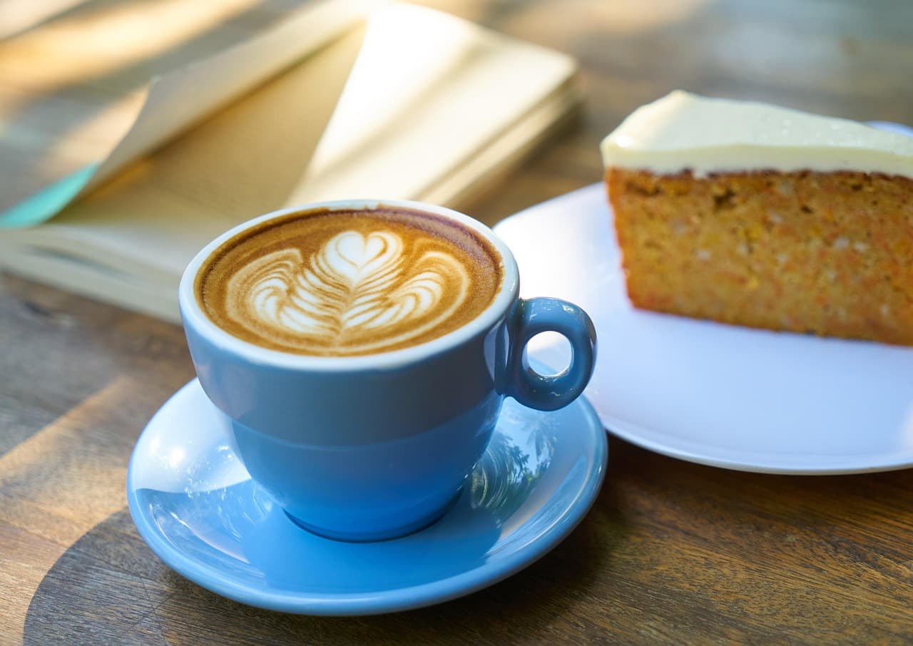 A cappuccino with latte art in a blue cup, a slice of sponge cake behind