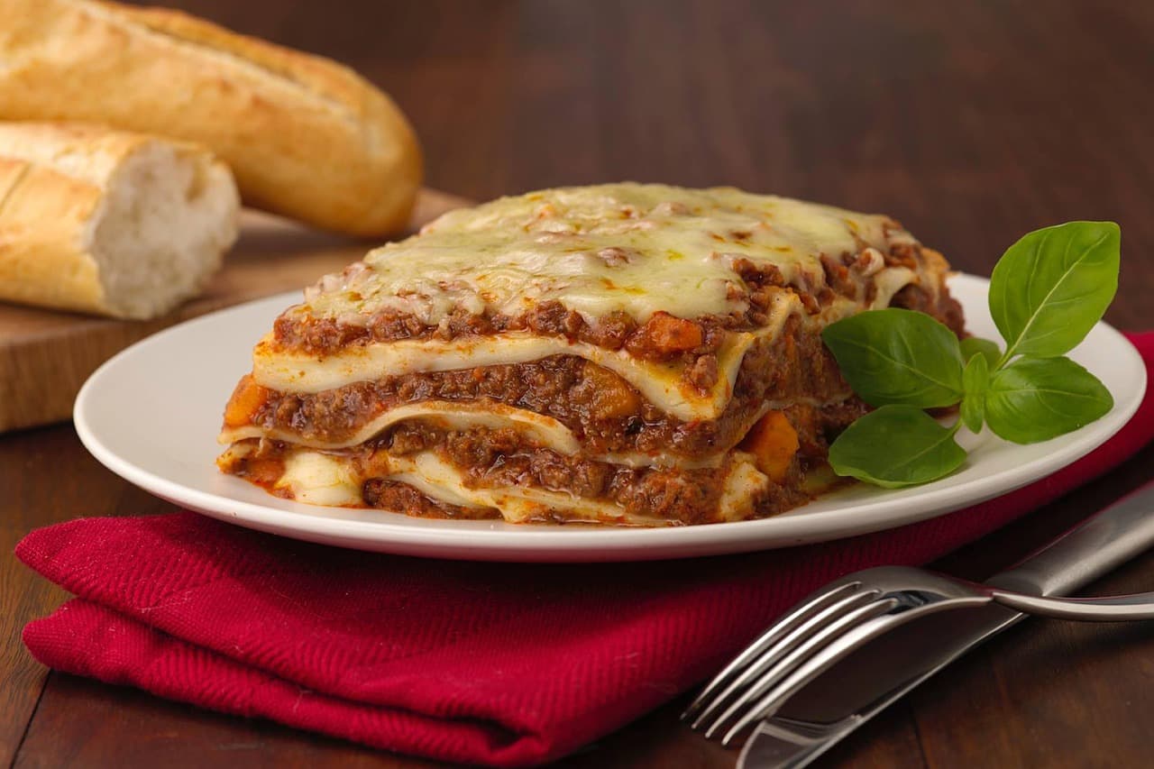 A slice of beef lasagne with basil on a white plate, bread beside it